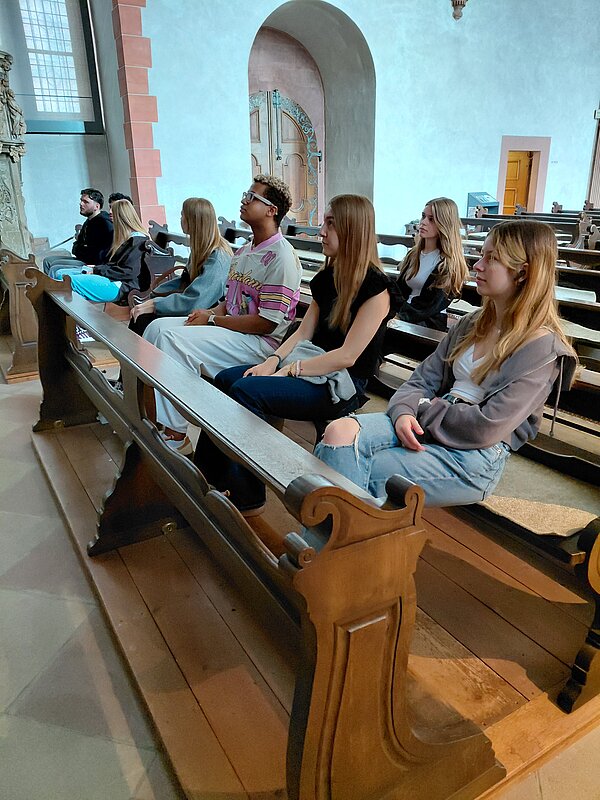 Eintauchen in die Geschichte Aschaffenburgs Mehrere junge Erwachsene sitzen aufmerksam nebeneinander auf einer Holzbank in einer Kirche. Die Atmosphäre ist ruhig und konzentriert.