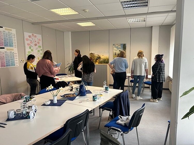 Eine Gruppe von Teilnehmenden steht im Raum und betrachtet die Materialien an der Wand. Der Raum ist mit langen Tischen und blauen Stühlen ausgestattet, auf denen Tassen, Krüge und Pflanzen stehen. An den Wänden hängen Poster und ein Landschaftsbild.