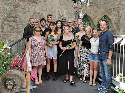 Eine Gruppe von jungen Menschen und einigen Lehrkräften in sommerlicher Kleidung stehen vor einer alten Burgmauer mit Fenstern. Die Auszubildenden halten Sonnenblumen in der Hand, einige Personen haben Sonnenbrillen auf. Oleander ragt von beiden Seiten leicht in das Bild hinein, vorne links ist ein Stein gemeißelter Löwenkopf zu erkennen.
