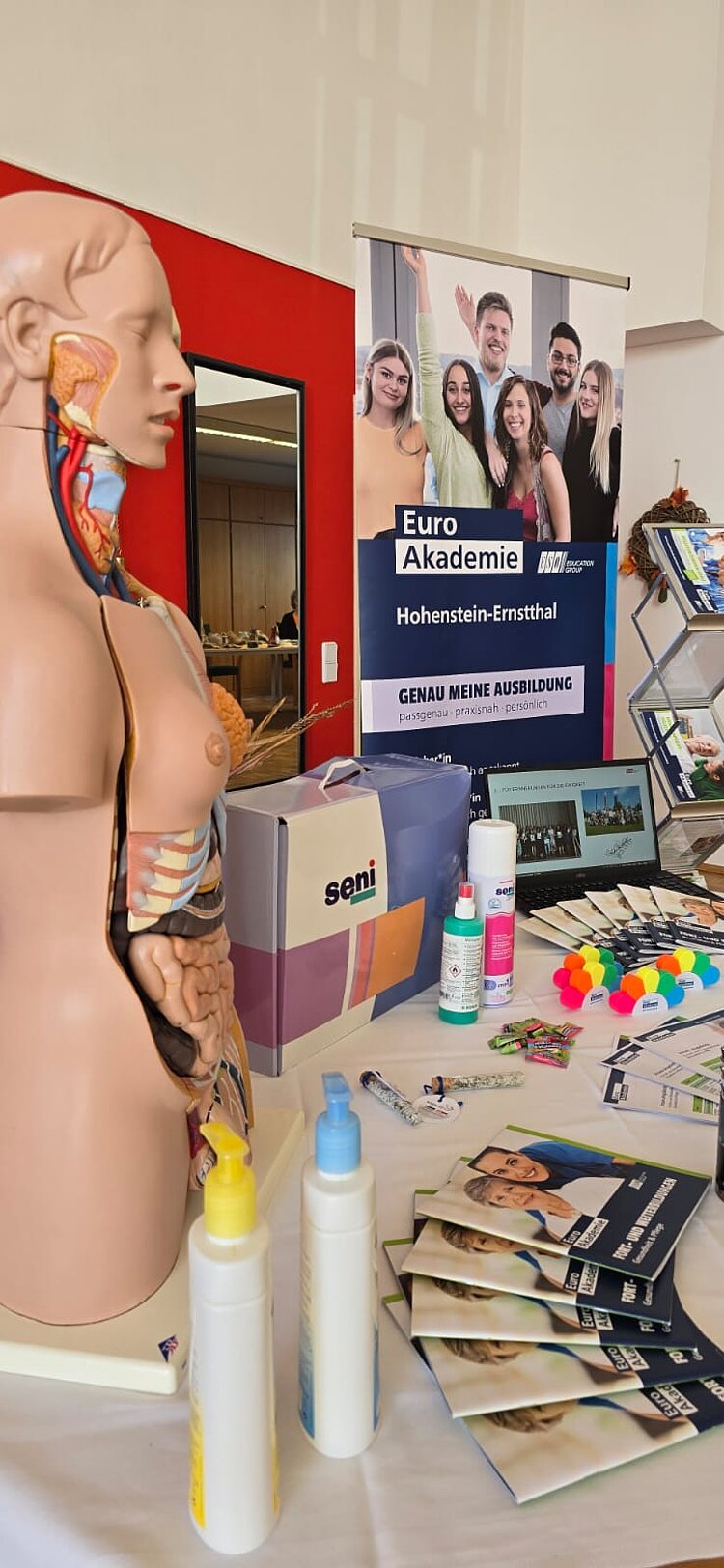 Auf einem Messetisch steht der Kunsstoff-Torso einer Frau, der teilweise den Blick auf die Organe sowie Adern und Venen freigibt. Außerdem liegt auf dem Tisch eine Menge Infomaterial und Messegeschenke. Hinter dem Tisch ist ein Roll-up der Euro Akademie Hohenstein-Ernstthal aufgestellt.