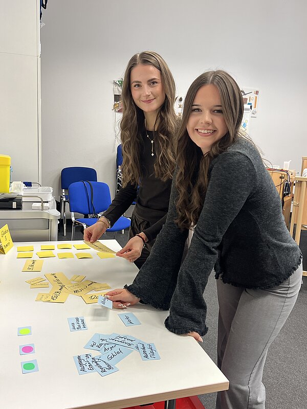 Zwei junge Frauen stehen lächelnd an einem Tisch und ordnen bunte Haftnotizen mit Stichworten für eine Gruppenarbeit.