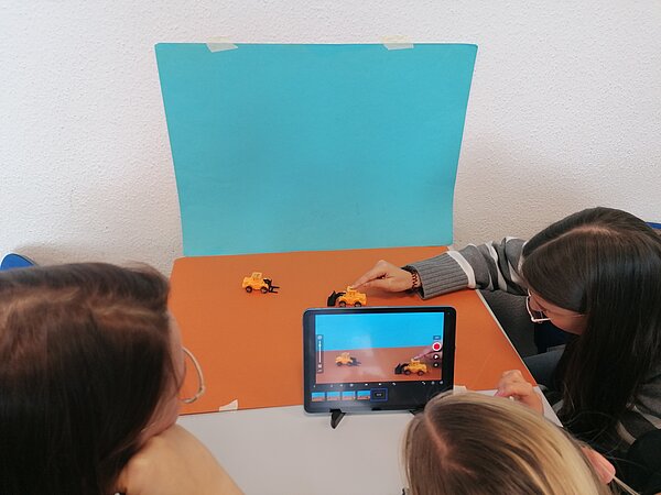 MeKoSax-Projekt „Trickfilmproduktion“ Drei Schülerinnen filmen drei kleine, gelbe Spielzeugbagger, welches auf einem hellbraunen Stück Papier stehen und vor einem hellblauen Hintergrund.