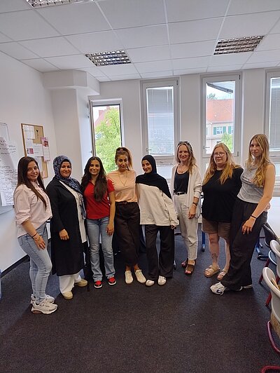 Gruppenfoto von acht Kursteilnehmenden in einem hellen Unterrichtsraum mit Fenstern, Whiteboard und Stühlen im Hintergrund.
