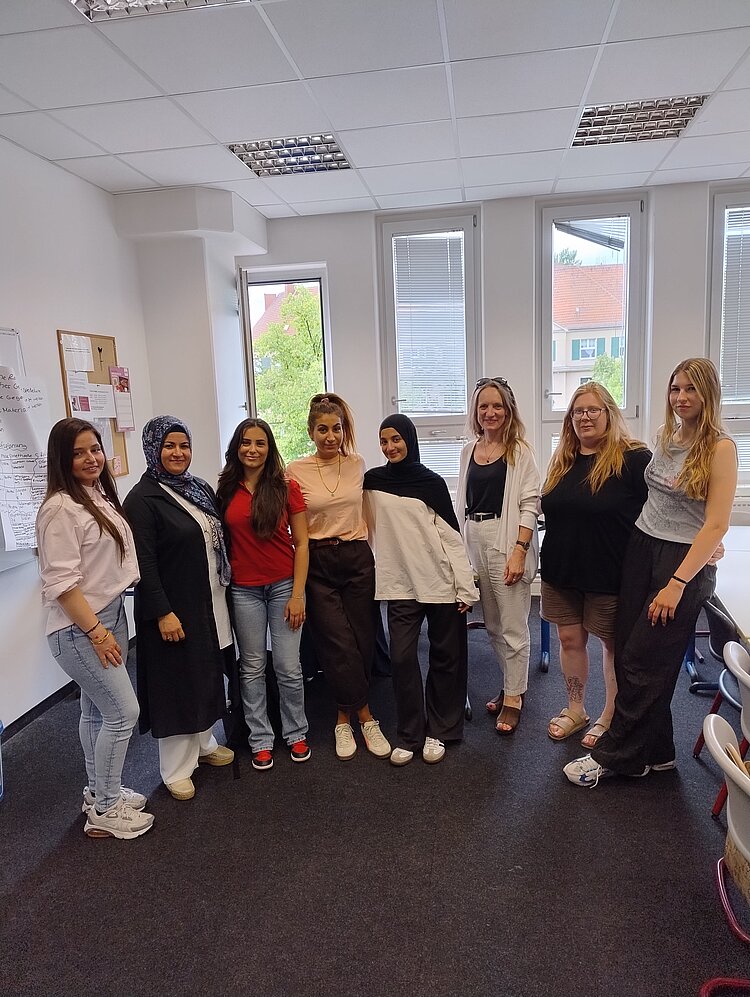 Gruppenfoto von acht Kursteilnehmenden in einem hellen Unterrichtsraum mit Fenstern, Whiteboard und Stühlen im Hintergrund.
