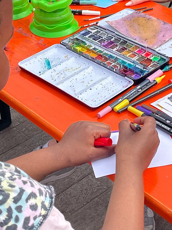Eine Person gestaltet an einem orangefarbenen Tisch ein Blatt Papier mit Stiften; daneben liegen Wasserfarben, Pinsel und weiteres Kreativmaterial.