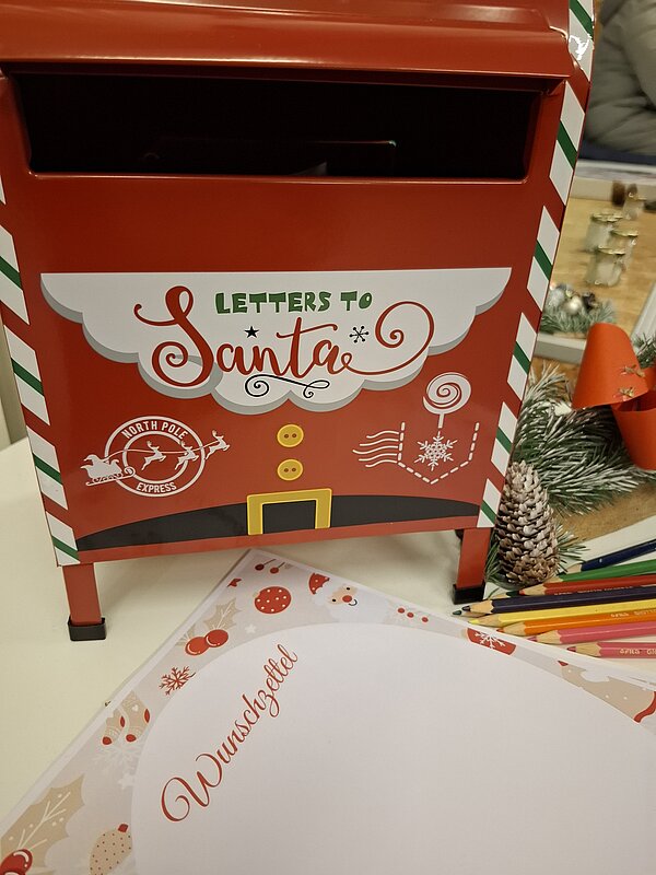 Roter Briefkasten mit der Aufschrift „Letters to Santa“ steht auf einem Tisch, davor liegt ein weihnachtlich gestalteter „Wunschzettel“ und bunte Stifte zum Schreiben der Wünsche.