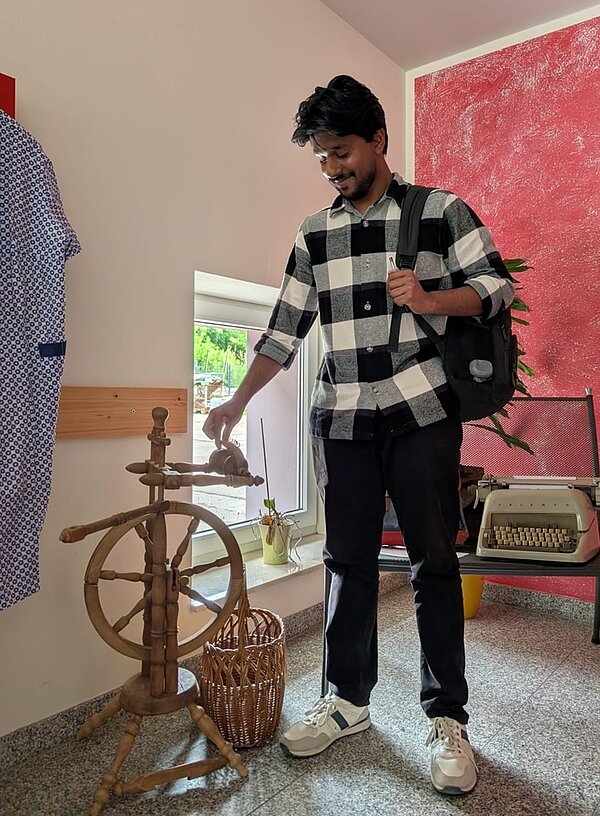 Ein Auszubildender mit kariertem Hemd und dunklem Rucksack steht in im Ausstellungsraum mit roter Wand und grauem Boden. Er hält eine Hand an einem hölzernen Spinnrad. Daneben steht ein Korb. Im Hintergrund befindet sich eine Schreibmaschine auf einem Tisch und ein Hemd hängt an der Wand.
