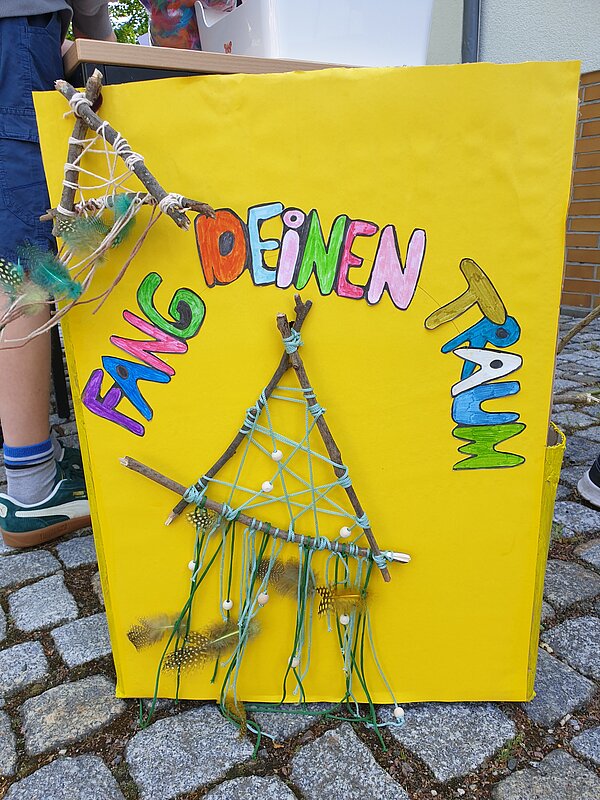 Ein erlebnisreicher Vormittag voller Spiel, Kreativität und Gemeinschaft Gelbes Schild mit der Aufschrift „Fang deinen Traum“ und kleinen, selbstgebastelten Traumfängern aus Ästen und Federn.