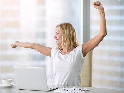 Die Szene zeigt eine Person mit erhobenen Armen vor einem Laptop auf einem Schreibtisch. Neben dem Laptop stehen eine Kaffeetasse und eine Brille. Das helle Zimmer hat große Fenster, die viel Tageslicht hereinlassen. Die Person trägt ein weißes Hemd und sitzt auf einem Bürostuhl.