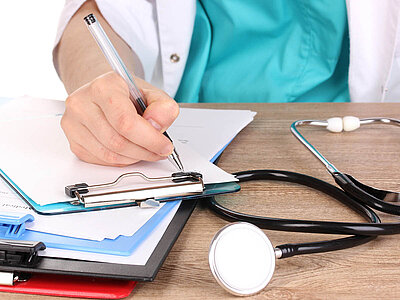 Ein Arzt schreibt auf einem Klemmbrett, während ein Stethoskop auf dem Tisch liegt. Professionelle Gesundheitsdokumentation.