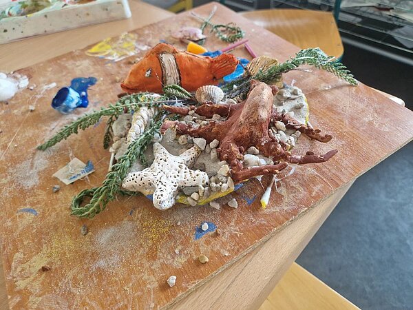 Ein handgefertigtes Tonrelief mit einem Unterwasser-Motiv. Das Relief zeigt einen Clown-Anemonenfisch, einen Tintenfisch, Algen, Muscheln und Steine, die auf einer Holzplatte arrangiert sind. Die Materialien wirken natürlich und detailliert, was die Vielfalt des Meeresbodens darstellen soll.