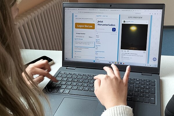 Eine Schülerin mit langen, hellen Haaren sitzt an einem Tisch und arbeitet an einem aufgeklappten Laptop. Die Hände befinden sich auf der Tastatur, während auf dem Bildschirm mehrere geöffnete Fenster sichtbar sind. Links ist eine Webseite mit dem Titel 'Legen Sie Los' und einem Button zu sehen, daneben ein weiteres Fenster mit einer Vorschau eines Social-Media-Posts. Im Hintergrund verläuft ein weißer Heizkörper entlang der Wand, der Raum ist hell und wirkt modern. Das Bild entstand im Rahmen des Projektes "Werde zum Faktenchecker" und zeigt eine typische Arbeitssituation beim digitalen Faktenchecken.