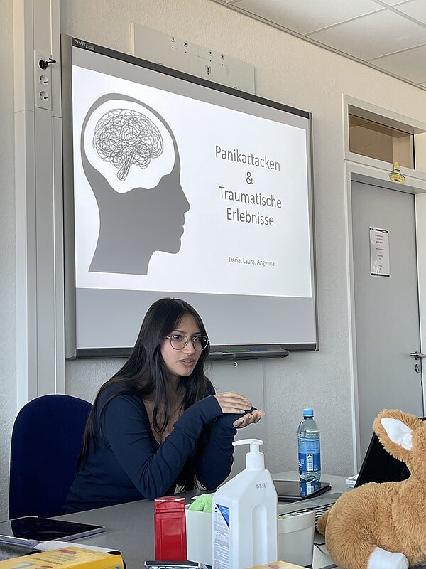 Eine Präsentationsfolie mit einer schwarz-weißen Linienzeichnung eines menschlichen Kopfes und eines Gehirns ist auf einer Leinwand zu sehen. Der Titel der Folie lautet: 'Panikattacken & Traumatische Erlebnisse'. Darunter stehen die Namen 'Daria, Laura, Angelina'. Im Vordergrund befindet sich eine Auszubildende mit langen dunklen Haaren, die an einem Tisch sitzt und gestikuliert. Auf dem Tisch stehen eine Wasserflasche, ein Desinfektionsmittel, ein Plüschtier und weitere Gegenstände. Im Hintergrund sind eine Tür und ein Fenster sichtbar.