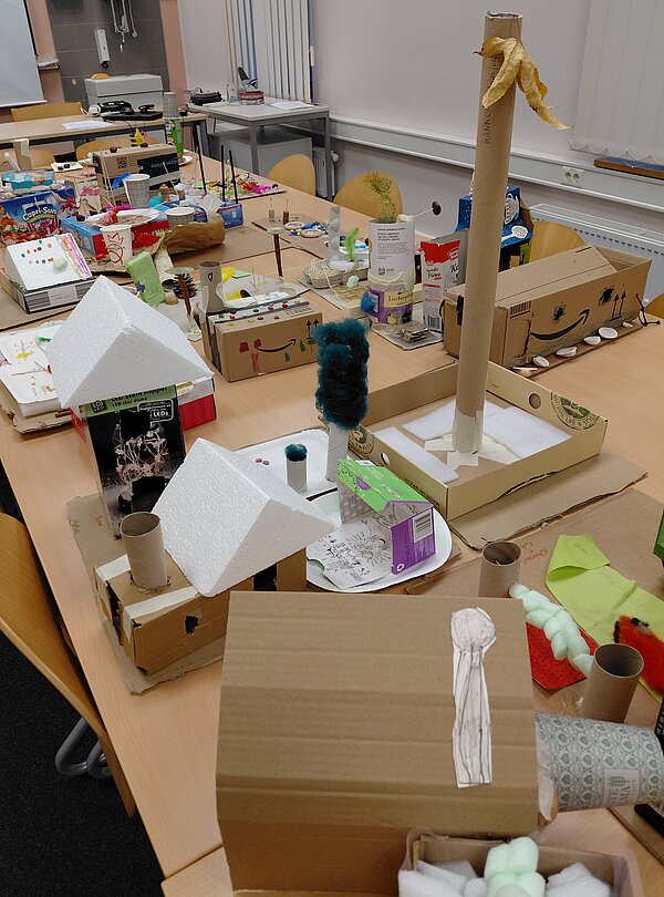 Ein langer Gruppentisch ist dicht gefüllt mit gebastelten Modellen aus Kartons, Styropor, Federn und Naturmaterialien; man erkennt kleine Häuser, Türme, Bäume und andere abstrakte Formen, die eine bunte Fantasielandschaft bilden.