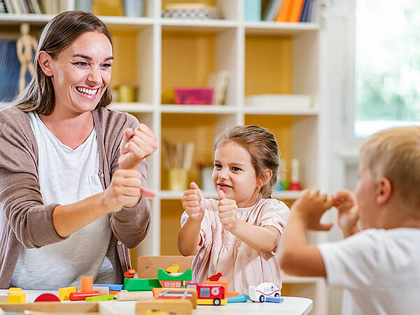 Eine Erwachsene und zwei Kinder spielen mit bunten Bauklötzen an einem Tisch vor einem Bücherregal. Die Erwachsene hält ein Spielzeugstück und gestikuliert, als würde er den Kindern etwas erklären oder mit ihnen spielen. Die Szene spielt sich wahrscheinlich in einem Umfeld einer Kindertagesstätte ab und betont Lern- oder Spielaktivitäten.