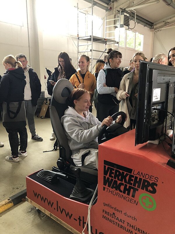 Ereignisreicher Tag für die angehenden Erzieher*innen In einer Halle sitzt eine Schülerin am Simulator eines Autositzes. Sie hält das Lenkrad fest in der Hand, vor ihr befindet sich ein Bildschirm. Der rote Simulator trägt die Aufschrift "Landesverkehrswacht Thüringen" inklusive des grünen Logos. In der Halle tummeln sich noch weitere Schülerinnen und Schüler.