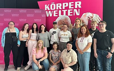 Eine Gruppe von Auszubildenden der Euro Akademie Halle sowie ihre Dozentin stehen und sitzen gemeinsam vor einer pinkfarbenen Wand mit dem Schriftzug „Körperwelten“. Alle blicken lächelnd in die Kamera.