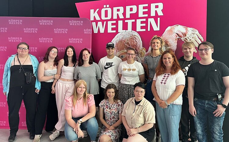 Echte Körper - echt faszinierend Eine Gruppe von Auszubildenden der Euro Akademie Halle sowie ihre Dozentin stehen und sitzen gemeinsam vor einer pinkfarbenen Wand mit dem Schriftzug „Körperwelten“. Alle blicken lächelnd in die Kamera.