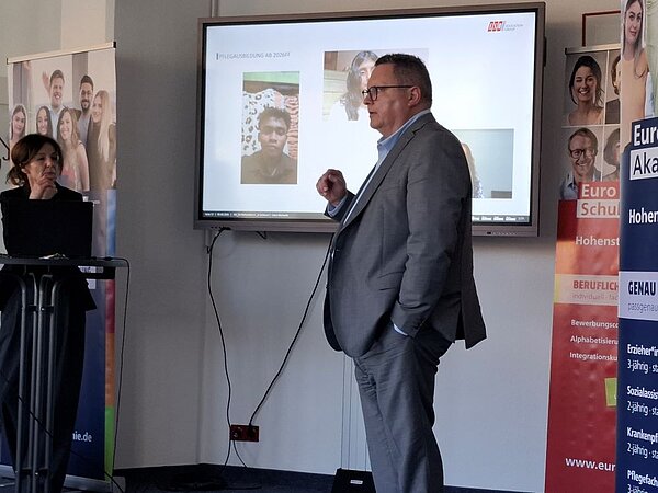 Ein Mann in einem Anzug steht vor einem Bildschirm und hält eine Präsentation. Auf dem Bildschirm sind Fotos von Pflegeausbildungen zu sehen. Daneben steht eine Frau an einem Pult.