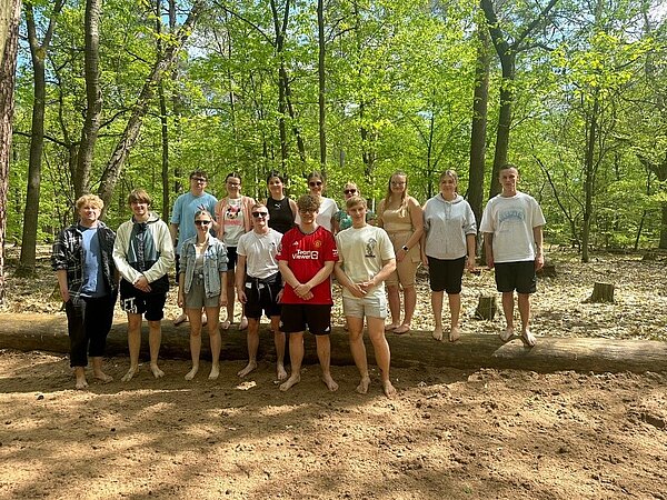Den Fläming mit allen Sinnen erleben Eine Gruppe von dreizehn jungen Menschen steht barfuß im Wald auf einem Baumstamm. Einige von ihnen tragen sportliche Kleidung, darunter ein rotes Liverpool-Trikot. Die Gruppe ist von hohen Bäumen und grünem Laub umgeben.