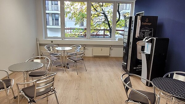 Aufenthaltsraum mit mehreren silbernen, runden Bistrotischen und passenden Metallstühlen. An der dunkelblau gestrichenen Wand steht eine große Snackmaschine.