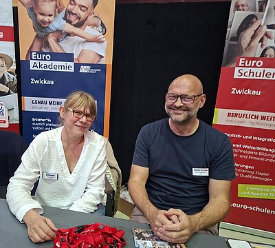 An einem Messestand sitzen eine lächelnde Frau und ein lächelnder Mann. Im Hintergrund stehen Roll-ups der Euro Akademie und Euro-Schulen Zwickau.