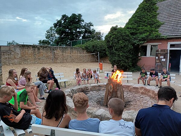 Sprachprojekte mit internationalem Flair Mädchen, Jungen und ihr Lehrkräfte sitzen in einem Kreis aus Bänken um ein Lagerfeuer.