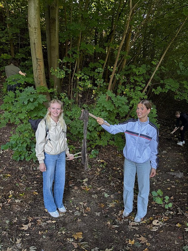 Zwei Schülerinnen der Euro Akademie Hohenstein-Ernstthal stehen im Wald. Eine der Schülerinnen präsentiert frisch gesammelten Müll in Form einer halb verrotteten Klamotte, die sie an ihrer Müllzange in die Kamera zeigt.