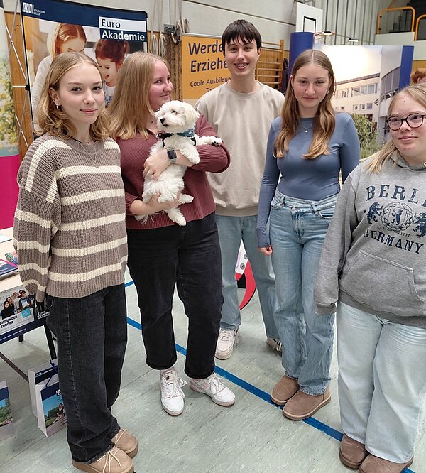 Auf dem Bild stehen fünf  Auszubildende der Euro Akademie Tauberbischofsheim gemeinsam vor dem Euro-Akademie-Messestandes auf der Ausbildungsmesse und posieren für ein Gruppenbild. Eine der Auszubildenden hat ein kleines, weißes Hündchen auf dem Arm.