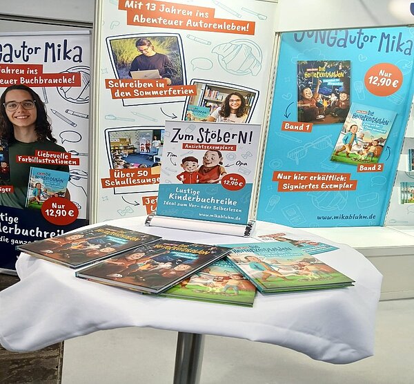 Die Leipziger Buchmesse – ein Erfahrungsbericht Ein Stehtisch, auf dem mehrere Bücher liegen. Dahinter Ausschnitte aus den Büchern in groß.