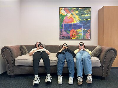 Drei Personen liegen auf einem Sofa und halten ihre Smartphones vor ihre Gesichter. Über dem Sofa hängt ein buntes Kunstwerk an der Wand.
