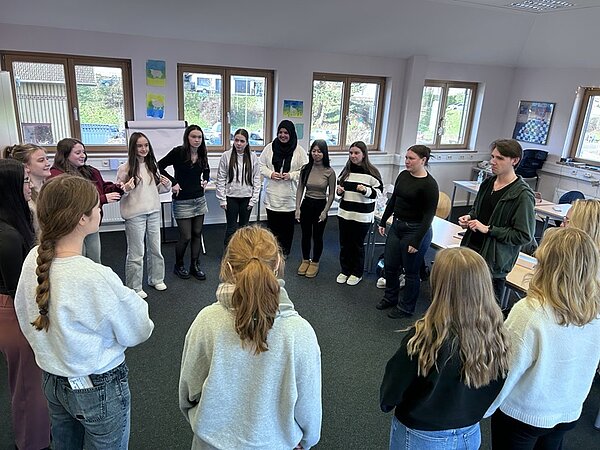In einem hellen Klassenraum der Euro Akademie Tauberbischofsheim stehen mehrere Schüler*innen in einem lockeren Kreis zusammen. Die Gruppe besteht aus Jugendlichen, die legere Kleidung wie Pullover, Jeans und Strickjacken tragen. Die meisten haben lange Haare, einige sind zu Zöpfen geflochten. Die Wände sind mit bunten Bildern und Zeichnungen dekoriert, und durch die großen Fenster fällt Tageslicht herein. Die Szene zeigt eine spontane Unterrichtsstunde, die im Rahmen des „aktiven“ Adventskalenders organisiert wurde.
