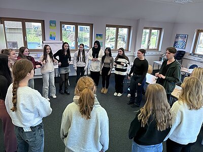 In einem hellen Klassenraum der Euro Akademie Tauberbischofsheim stehen mehrere Schüler*innen in einem lockeren Kreis zusammen. Die Gruppe besteht aus Jugendlichen, die legere Kleidung wie Pullover, Jeans und Strickjacken tragen. Die meisten haben lange Haare, einige sind zu Zöpfen geflochten. Die Wände sind mit bunten Bildern und Zeichnungen dekoriert, und durch die großen Fenster fällt Tageslicht herein. Die Szene zeigt eine spontane Unterrichtsstunde, die im Rahmen des „aktiven“ Adventskalenders organisiert wurde.