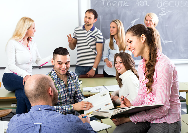 Personen unterschiedlichen Alters sitzen und stehen in einem Klassenzimmer. Sie halten teilweise geöffnete Bücher in der Hand und diskutieren. Auf der Tafel im Hintergrund steht: "Theme for discussion".