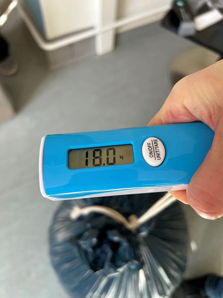 Sammelaktion: Kronkorken für den guten Zweck Eine Hand hält eine digitale Kofferwaage mit blauem Gehäuse. Das Display zeigt ein Gewicht von 18,0 kg an. Im Hintergrund ist ein schwarzer Müllsack zu sehen, der die gesammelten Kronkorken enthält.