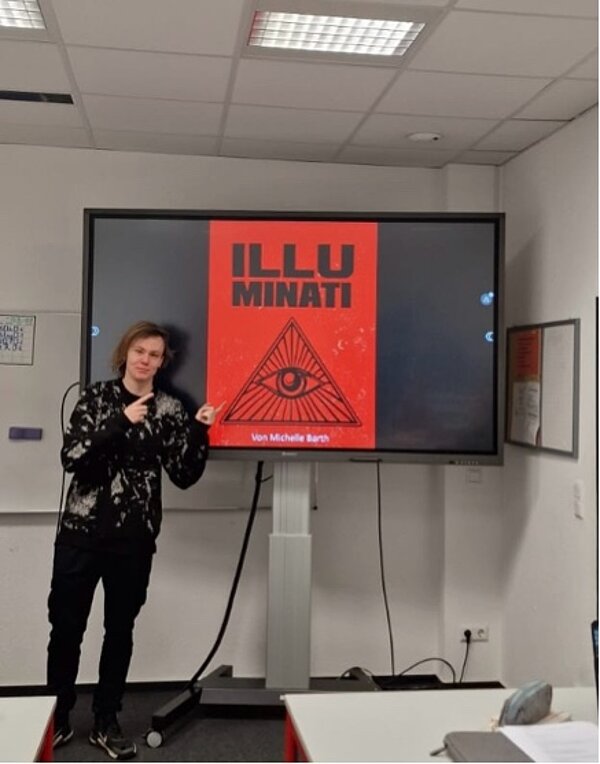 Ein Schüler steht vor einem Bildschirm mit einer roten Präsentationsfolie. In der Mitte ist das Symbol der Illuminaten (Auge in der Pyramide) zu sehen. Der Titel der Präsentation lautet „ILLUMINATI“, darunter steht „Von Michelle Barth“. Der Schüler zeigt mit beiden Händen auf die Folie.