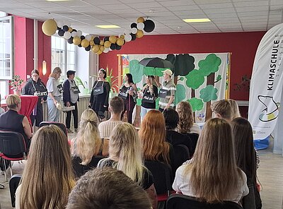 Mehrere Personen führen vor Publikum ein kleines Theaterstück auf. Im Hintergrund ist eine selbst gestaltete Waldkulisse zu sehen, der Raum ist mit schwarz-weißen und goldenen Luftballons dekoriert.