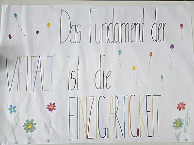 Ein Plakat mit dem bunten Schriftzug "Das Fundament der Vielfalt ist die Einzigartigkeit", umringt von gezeichneten Blumen und Luftballons.