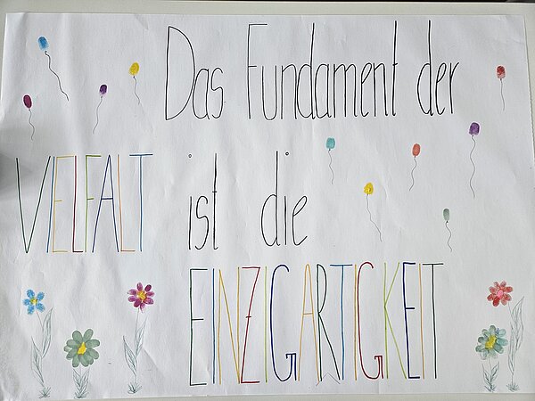 Projekt Vielfältigkeitsräder Ein Plakat mit dem bunten Schriftzug "Das Fundament der Vielfalt ist die Einzigartigkeit", umringt von gezeichneten Blumen und Luftballons.