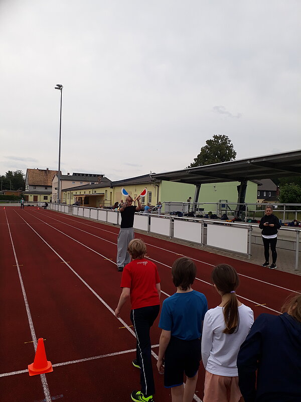 Unser Motto zum Sportfest: "Auf zu neuen Galaxien!" Kinder stehen auf einer roten Laufbahn in einer Reihe. Ein Mann vorne bereitet sich vor, ein Startsignal mit zwei Fahnen zu geben.