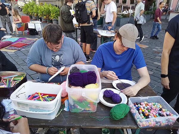 Zwei junge Erwachsene sitzen am Basteltisch und arbeiten mit bunter Wolle und Perlen, um Pappteller mit Garn zu gestalten.