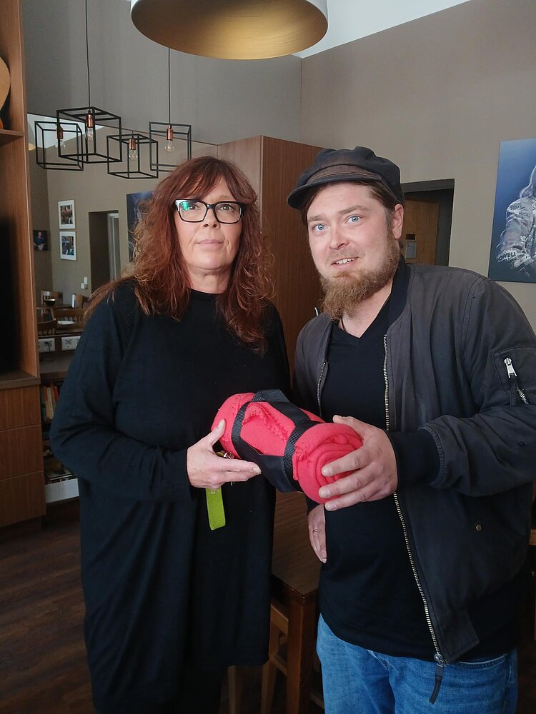 Eine Frau und ein Mann stehen in einem Café und halten gemeinsam eine aufgerollte rote Decke.