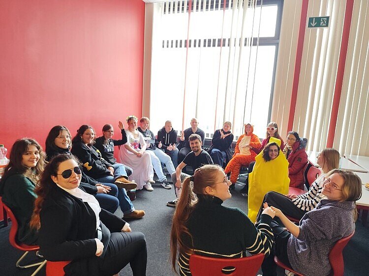 Helau: FSK und FAK feiern zusammen Fasching In einem Klassenzimmer sitzt eine große Gruppe Schüler*innen, die fast alle unterschiedlich verkleidet sind. Sie bilden einen Stuhlkreis und blicken in die Kamera.