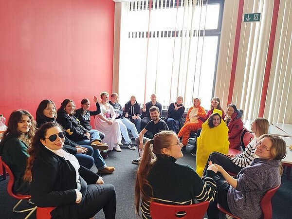 Helau: FSK und FAK feiern zusammen Fasching In einem Klassenzimmer sitzt eine große Gruppe Schüler*innen, die fast alle unterschiedlich verkleidet sind. Sie bilden einen Stuhlkreis und blicken in die Kamera.