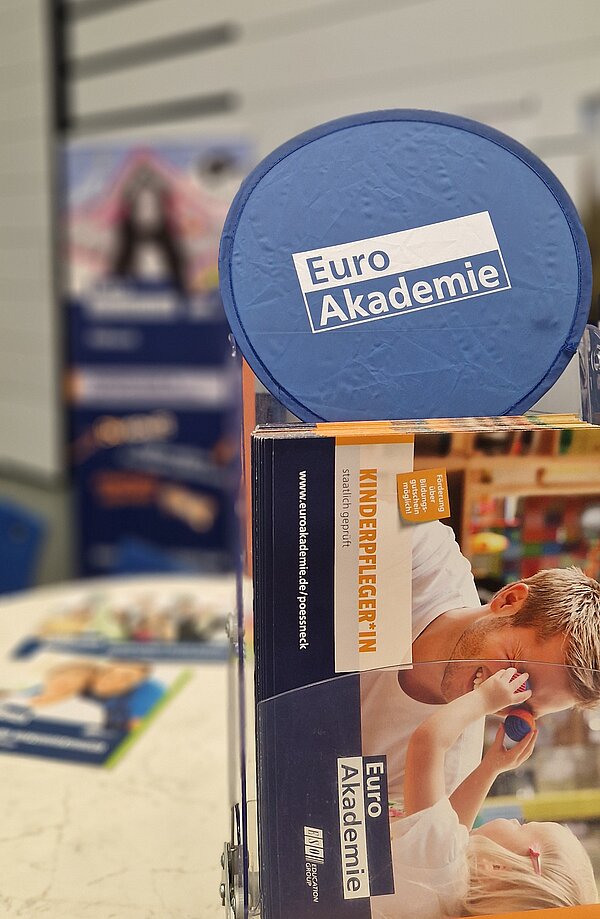 Prospektständer mit Flyern der Euro Akademie und blauem Werbeartikel im Vordergrund, Messestand unscharf im Hintergrund.