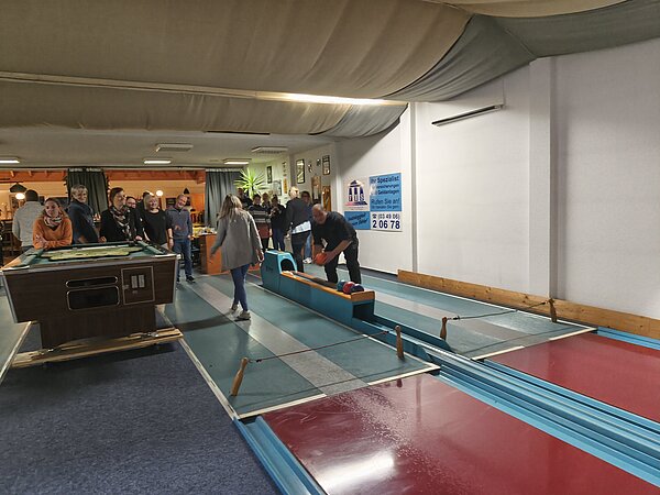 Kegelbahn: Gruppe schaut zu, während eine Person die Kugel ansetzt; im Vordergrund Billardtisch, im Hintergrund fröhliche Runde.