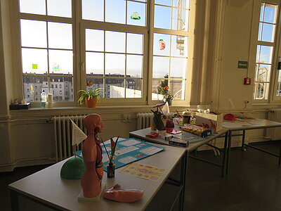 In einem Klassenraum stehen auf Tischen vor den Fenstern verschiedene Gegenstände, darunter ein Modell der menschlichen Organe, eine Animefigur aus Pappmaché, verschiedene Spiele und Gebäck.