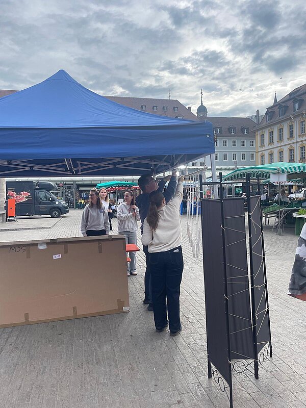 Mehrere Personen bauen auf einem Marktplatz einen blauen Pavillon für eine Aktion oder Veranstaltung auf.
