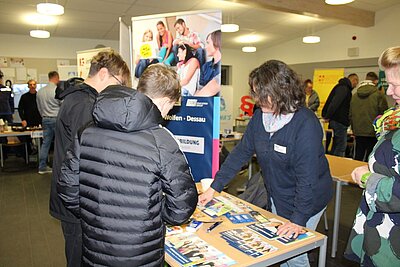 Eine lebhafte Informationsmesse in einem hellen Raum. Im Vordergrund steht eine Frau mit Namensschild an einem Tisch, der mit Broschüren und Flyern bedeckt ist. Sie erklärt zwei jungen Männern, die ihr den Rücken zukehren, etwas zu den Materialien. Beide Männer tragen dunkle Jacken. Im Hintergrund sind weitere Besucher*innen und Stände zu sehen, darunter ein Stand mit der Aufschrift 'Wolfen - Dessau' und 'Ausbildung'. Der Raum wirkt belebt und gut besucht.