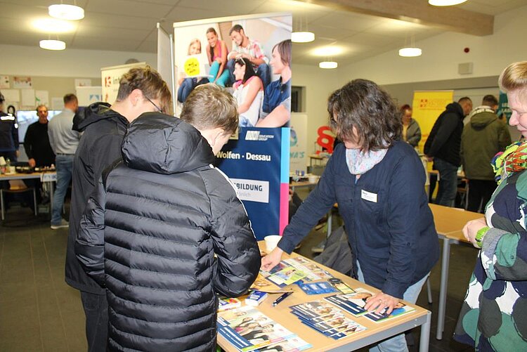 Eine lebhafte Informationsmesse in einem hellen Raum. Im Vordergrund steht eine Frau mit Namensschild an einem Tisch, der mit Broschüren und Flyern bedeckt ist. Sie erklärt zwei jungen Männern, die ihr den Rücken zukehren, etwas zu den Materialien. Beide Männer tragen dunkle Jacken. Im Hintergrund sind weitere Besucher*innen und Stände zu sehen, darunter ein Stand mit der Aufschrift 'Wolfen - Dessau' und 'Ausbildung'. Der Raum wirkt belebt und gut besucht.